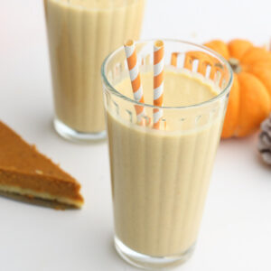 a glass of pumpkin pie milkshake with 2 straws, a piece of pie, small pumpkins and a pinecone